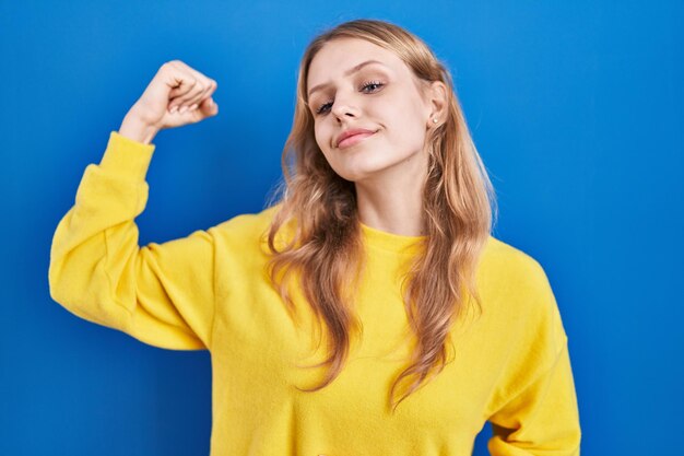 Mujer caucásica joven de pie sobre una persona fuerte de fondo azul que muestra músculo del brazo, segura y orgullosa del poder