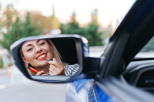 Mujer caucásica joven que aplica el lápiz labial que mira el reflejo en el espejo del coche.