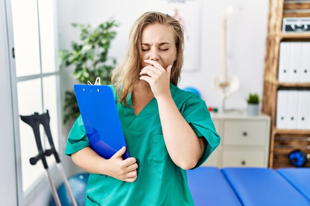 Mujer caucásica joven que trabaja en la clínica de recuperación del dolor aburrida bostezando cansada cubriendo la boca con la mano inquieta y somnolencia