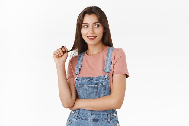 Mujer caucásica joven seductora femenina en camiseta de overol tonta hebra de cabello ondulado en el dedo y mirando a la derecha con expresión intrigada y entusiasta mirando el fondo blanco de la chica caliente