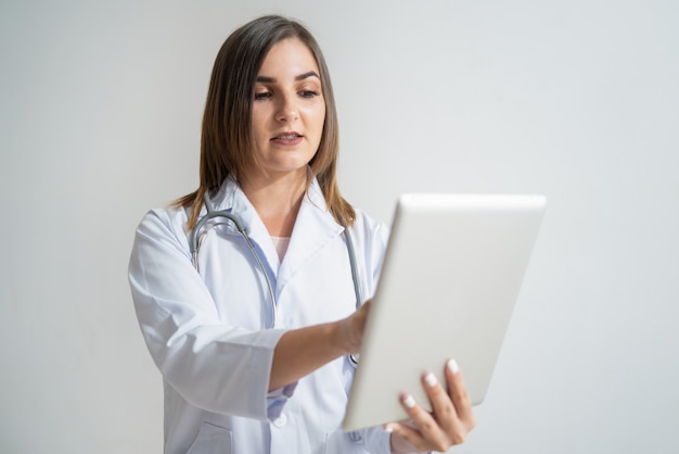 Mujer caucásica joven seria en la capa del laboratorio que mira la tableta