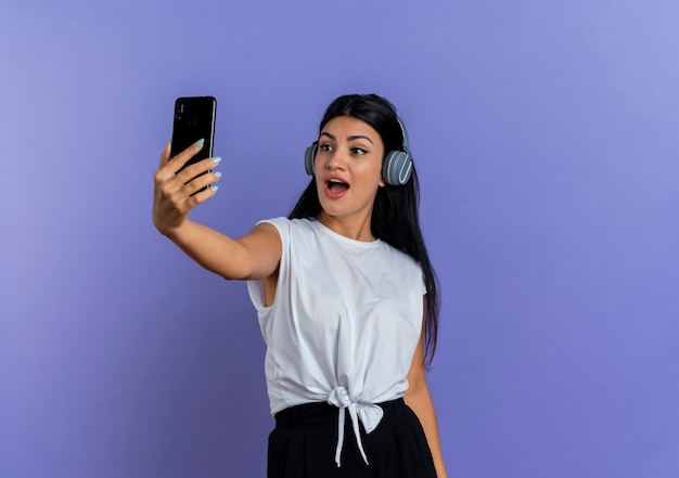 Mujer caucásica joven sorprendida en auriculares mira teléfono