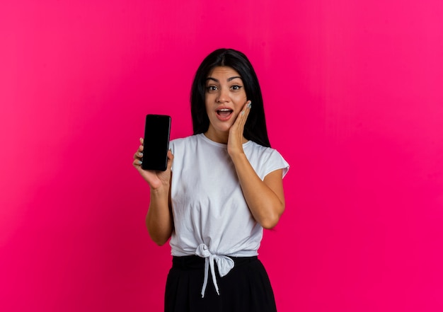 Mujer caucásica joven sorprendida pone la mano en la cara y sostiene el teléfono