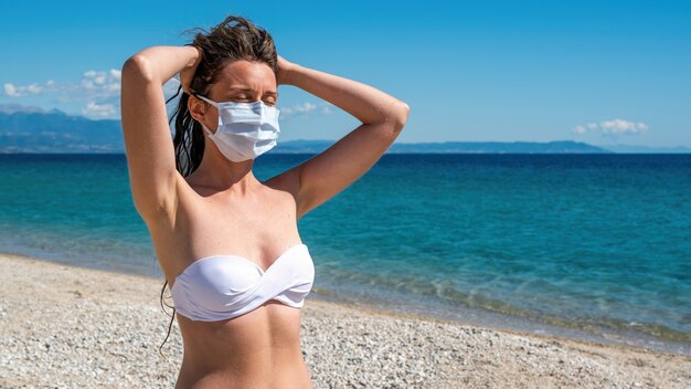 Mujer caucásica en máscara médica blanca con las manos levantadas a la cabeza y los ojos cerrados en traje de baño en una playa en Asprovalta, Grecia