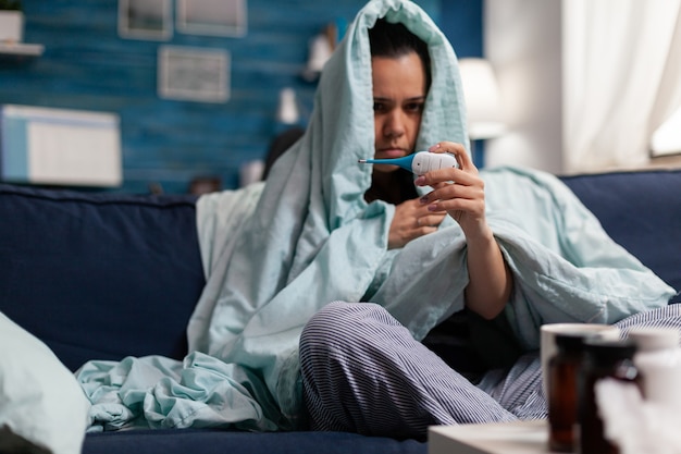 Mujer caucásica medir la temperatura enferma en casa con un termómetro. Persona que se siente enferma por el frío, que controla la fiebre y los síntomas de la enfermedad de la gripe. Adulto en reposo con dolor de cabeza