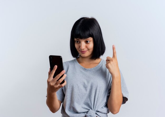 Mujer caucásica morena joven sorprendida mira el teléfono y señala hacia arriba