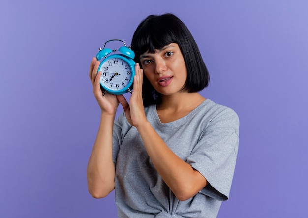 Mujer caucásica morena joven sorprendida sostiene el reloj de alarma aislado sobre fondo púrpura con espacio de copia