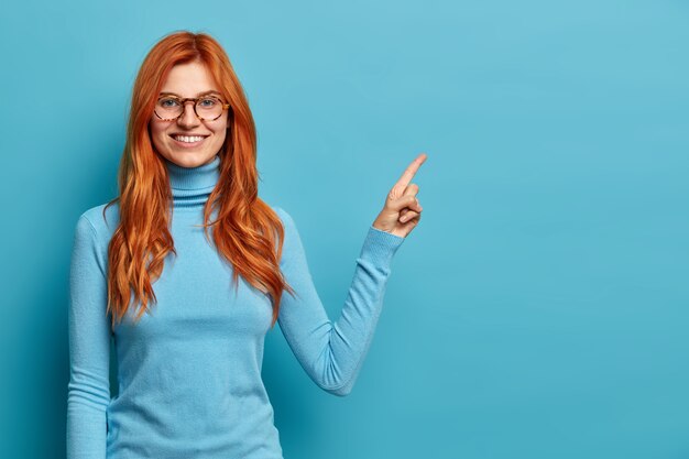 Mujer caucásica pelirroja de aspecto agradable positivo con puntos de sonrisa con dientes en el espacio de la copia en la esquina superior derecha.