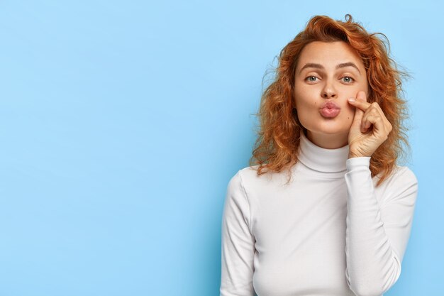 Mujer caucásica pelirroja coqueta mantiene los labios doblados, da mwah romántico tierno