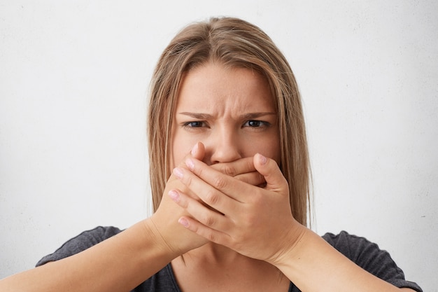 Foto gratuita mujer caucásica de pelo rubio con mirada enojada frunciendo el ceño y cubriendo la boca con las manos tratando de sujetar la lengua. mujer elegante tratando de guardar silencio y no contar secretos