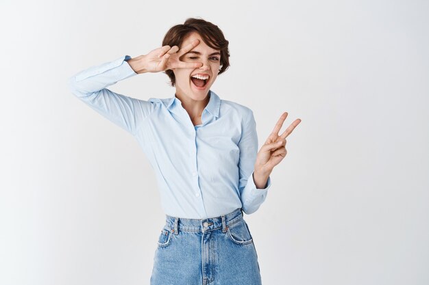 Mujer caucásica positiva y feliz sonriendo, mostrando signos de paz y divirtiéndose, de pie optimista en la pared blanca