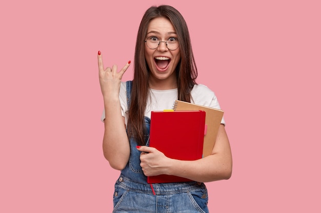 Mujer caucásica positiva hace gesto de rock n roll lleva libros de texto se siente llena de alegría y genial