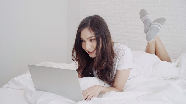 Mujer caucásica de la raza asiática hermosa de la mezcla que usa el ordenador o el ordenador portátil mientras que miente en la cama en su dormitorio.