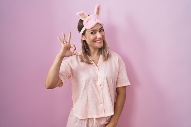 Mujer caucásica rubia con antifaz para dormir y pijama sonriendo positivamente haciendo el signo de ok con la mano y los dedos expresión exitosa