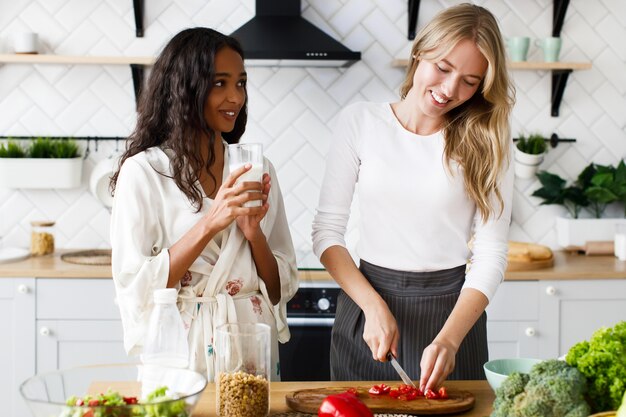 La mujer caucásica rubia sonrió está cortando pimienta y la mujer mulata morena en ropa de dormir está bebiendo leche y mirándola en la cocina moderna blanca