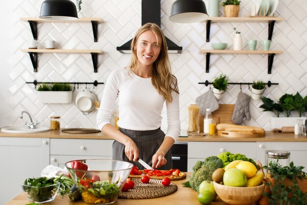 La mujer caucásica rubia sonrió está cortando el pimiento rojo en la cocina moderna en la mesa llena de frutas y verduras frescas