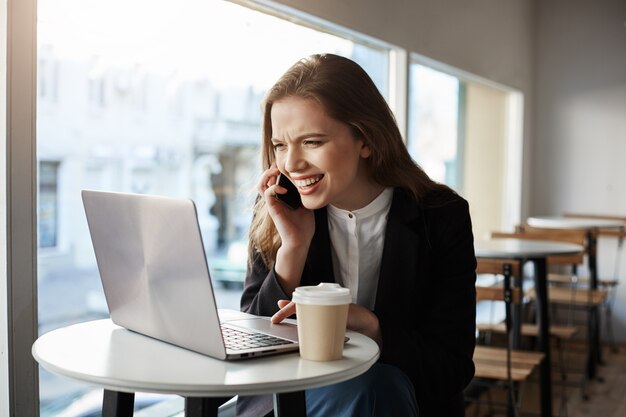 Mujer caucásica sentada en la cafetería, tomando café, hablando por teléfono inteligente, mirando la pantalla del portátil con una amplia sonrisa