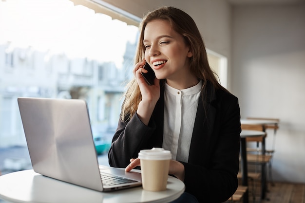 Mujer caucásica sentada en la cafetería, tomando café, hablando por teléfono inteligente, mirando la pantalla del portátil con una amplia sonrisa