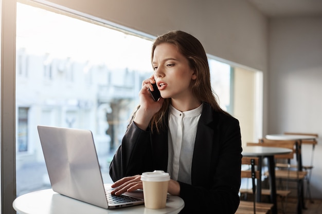 Mujer caucásica sentado en la cafetería con laptop, tomando café, hablando por teléfono inteligente