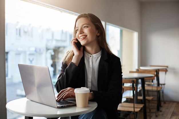 Mujer caucásica sentado en la cafetería con laptop, tomando café, hablando por teléfono inteligente