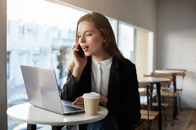 Mujer caucásica sentado en la cafetería con laptop, tomando café, hablando por teléfono inteligente