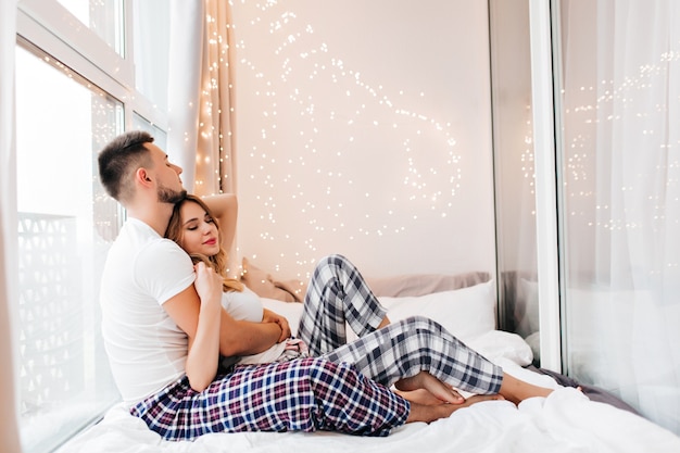 Mujer caucásica soñolienta posando en la cama por la mañana. Chica cansada en pijama junto a su novio.