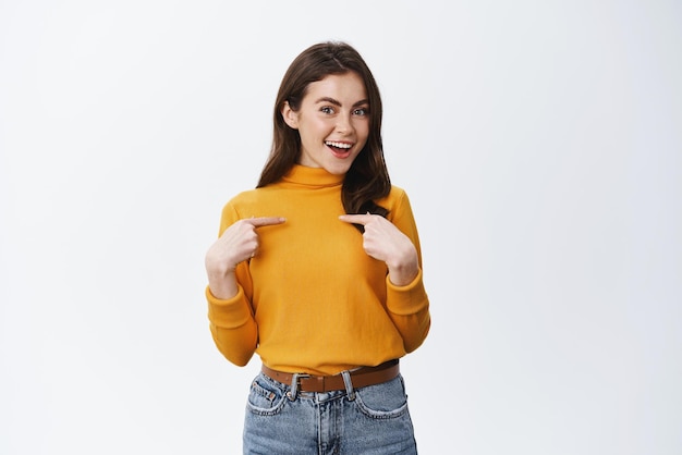 Foto gratuita una mujer caucásica sonriente apuntándose a sí misma y autopromocionándose presenta o elogia sus propios logros de pie sobre fondo blanco confiada