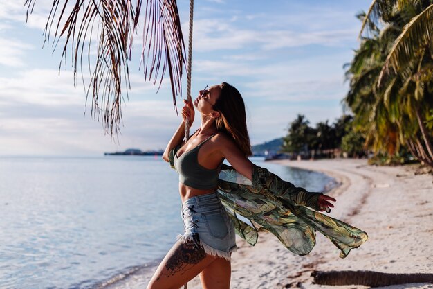 Mujer caucásica tatuada en pantalones cortos de mezclilla y top verde de moda en la playa