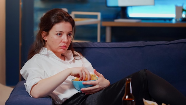 Mujer caucásica tendido en el sofá y comer bocadillos mientras ve la televisión en casa. Adulto joven con tazón de patatas fritas en la mano y relajarse en el sofá después del trabajo mirando la televisión en la sala de estar.