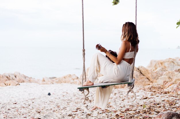 Mujer caucásica tranquila feliz joven romántica con el ukelele en la playa rocosa tropical al atardecer