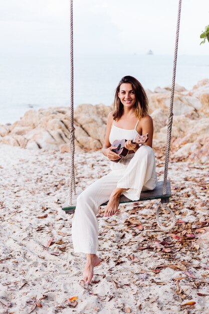 Mujer caucásica tranquila feliz joven romántica con el ukelele en la playa rocosa tropical al atardecer