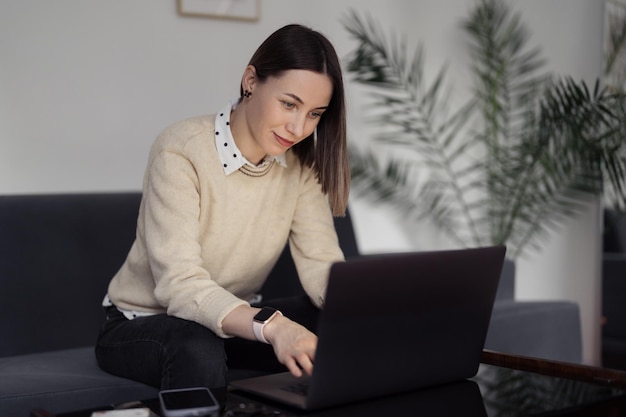 Mujer caucásica usando una computadora portátil mientras se sienta en el sofá en casa en la sala de estar de la noche