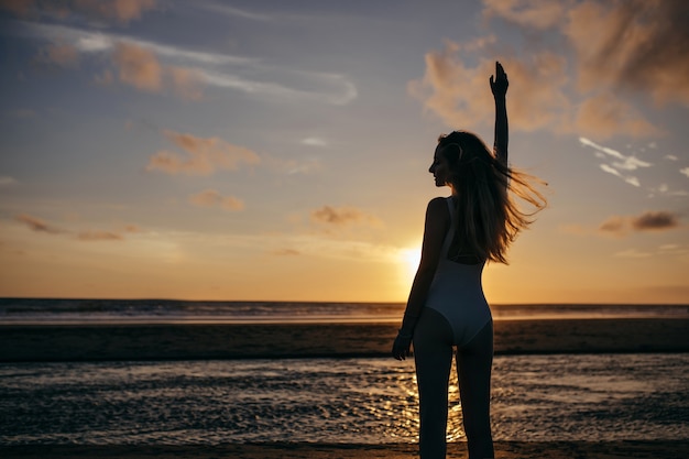 mujer caucásica viste traje de baño blanco en vacaciones. Señora joven despreocupada disfrutando de la noche en el océano y mirando la hermosa puesta de sol.