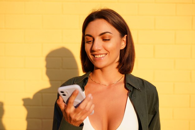 Mujer caucásica vistiendo camiseta al atardecer en la pared de ladrillo amarillo exterior mirada positiva en la pantalla del teléfono móvil con una sonrisa