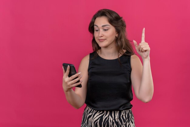 Mujer caucásica vistiendo camiseta negra sosteniendo un teléfono y apunta hacia arriba en la pared rosa