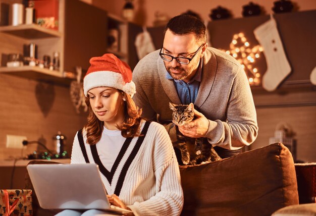 Mujer celebrando la Nochebuena en casa con gato. Pareja casada usando una computadora portátil.