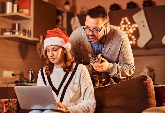 Mujer celebrando la Nochebuena en casa con gato. Pareja casada usando una computadora portátil.