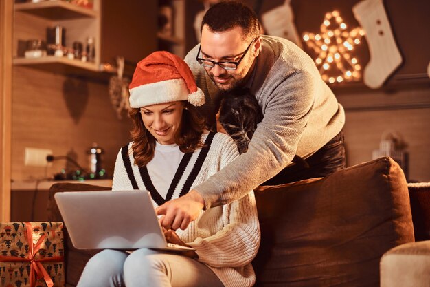 Mujer celebrando la Nochebuena en casa con gato. Pareja casada usando una computadora portátil.