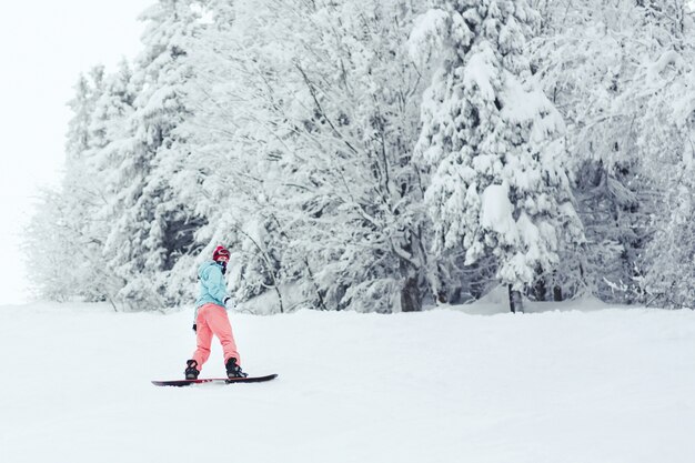 La mujer en la chaqueta de esquí azul y los pantalones rosados ​​se coloca en el snowboard en algún lugar en bosque del invierno