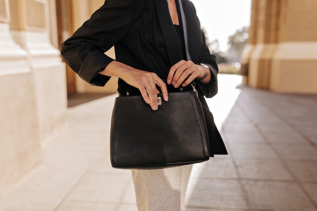Mujer de chaqueta negra y pantalón blanco con bolso oscuro afuera. mujer en ropa moderna posando con elegante bolso al aire libre.