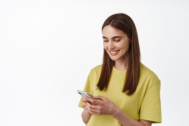 mujer chateando, enviando mensajes de texto en el teléfono, usando la aplicación del teléfono inteligente, leyendo la pantalla y sonriendo, de pie en blanco