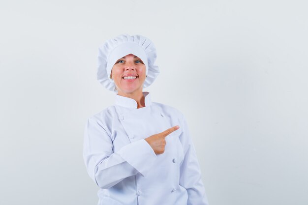 Mujer chef apuntando a la esquina superior derecha con uniforme blanco y mirando confiado