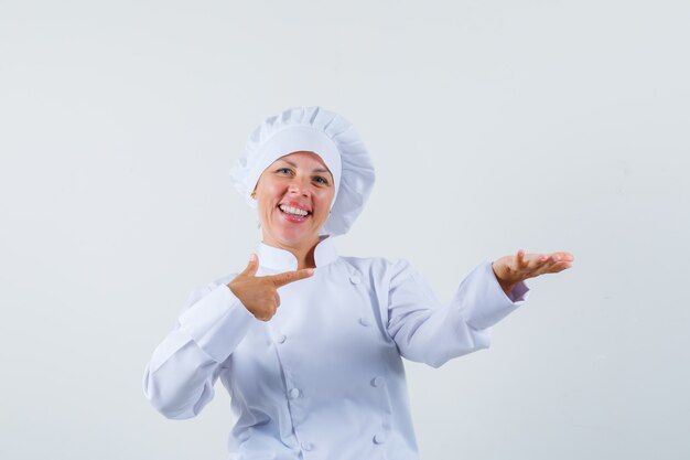 mujer chef apuntando a su mano izquierda mientras sostiene algo en uniforme blanco y se ve optimista.