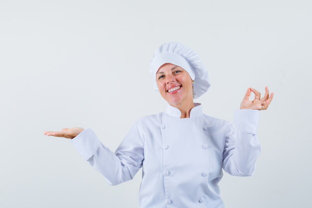 mujer chef mostrando gesto de ganador mientras extiende la palma a un lado en uniforme blanco y parece complacida.