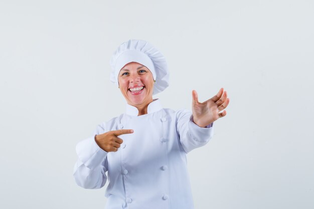 Mujer chef posando como apuntando al teléfono en uniforme blanco y mirando alegre