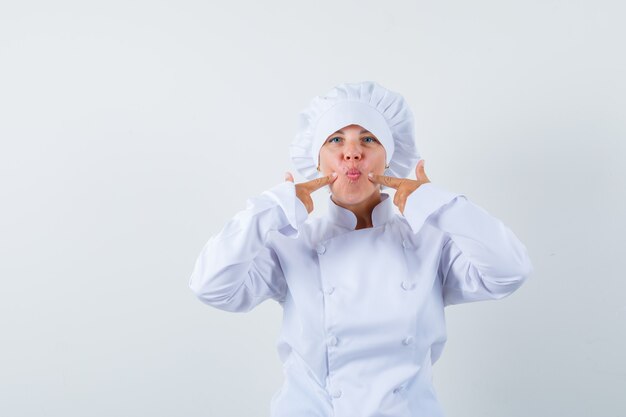 Mujer chef presionando los dedos en las mejillas en uniforme blanco y mirando divertido