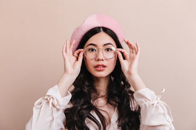 Mujer China curiosa posando en vasos. Mujer asiática de moda en boina aislada sobre fondo beige.