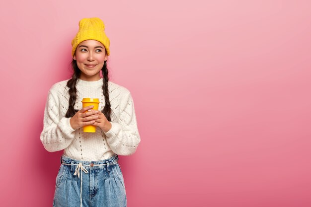 Mujer china de ensueño bebe café para llevar, se levanta pensativamente contra el fondo rosa