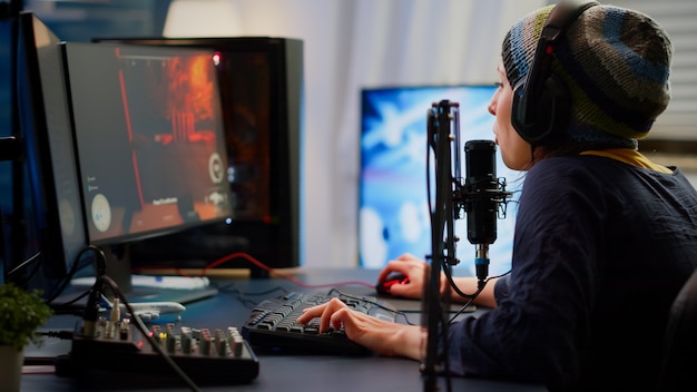 Mujer cibernética profesional hablando en el micrófono profesional de transmisión en el estudio de juegos en casa con chat de flujo abierto. Jugador que participa en un torneo en línea utilizando una potente computadora personal con RGB y auriculares.