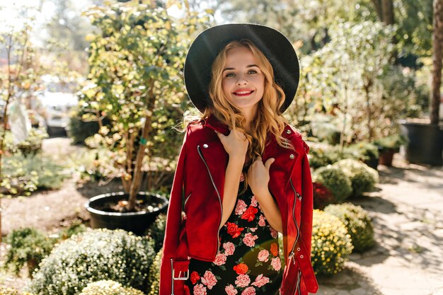 Mujer ciega con labios rojos se pone chaqueta de cuero roja. mujer con sombrero negro posando en el jardín.